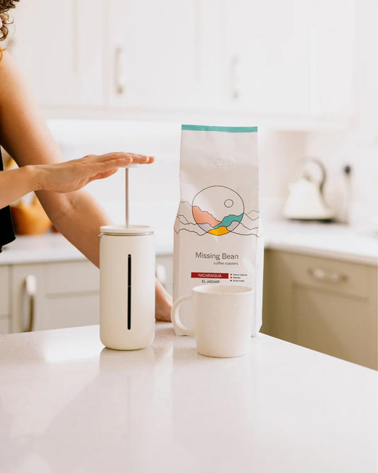 Person pressing down a French press beside a coffee bag labeled "Missing Bean Coffee Roasters, Nicaragua El Jaguar" and a white mug on a kitchen counter.