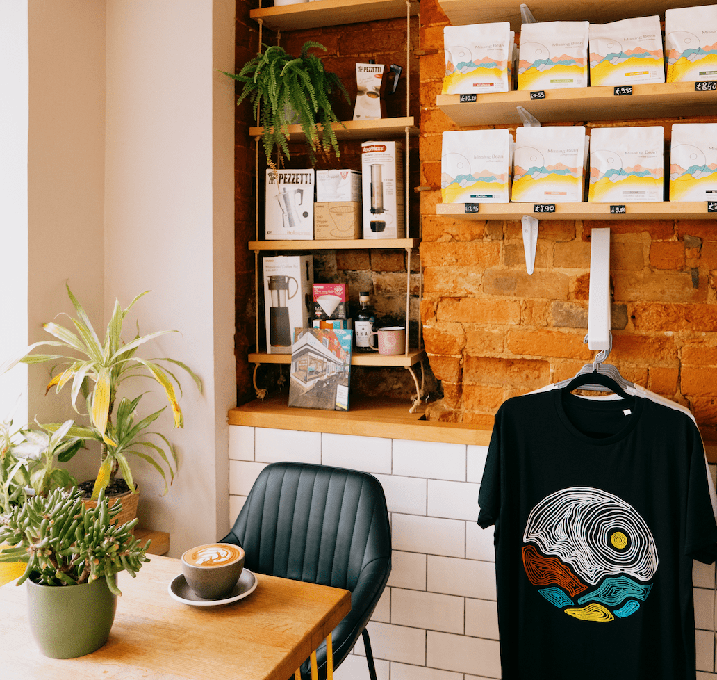 Scene inside Missing Bean Coffee Shop in Banbury, Oxford. There is a coffee on wooden table and shelves containing coffee beans and merchandise for sale.