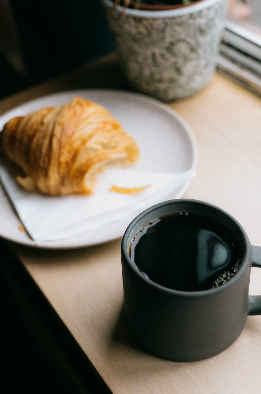 A black ceramic mug filled with coffee sits on a wooden table next to a white plate with a croissant on it. A green and white patterned plant pot is partially visible in the background. The scene is cozy and inviting, bathed in soft natural light.