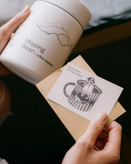 Person holding a Missing Bean coffee container and a gift subscription envelope with an illustration.