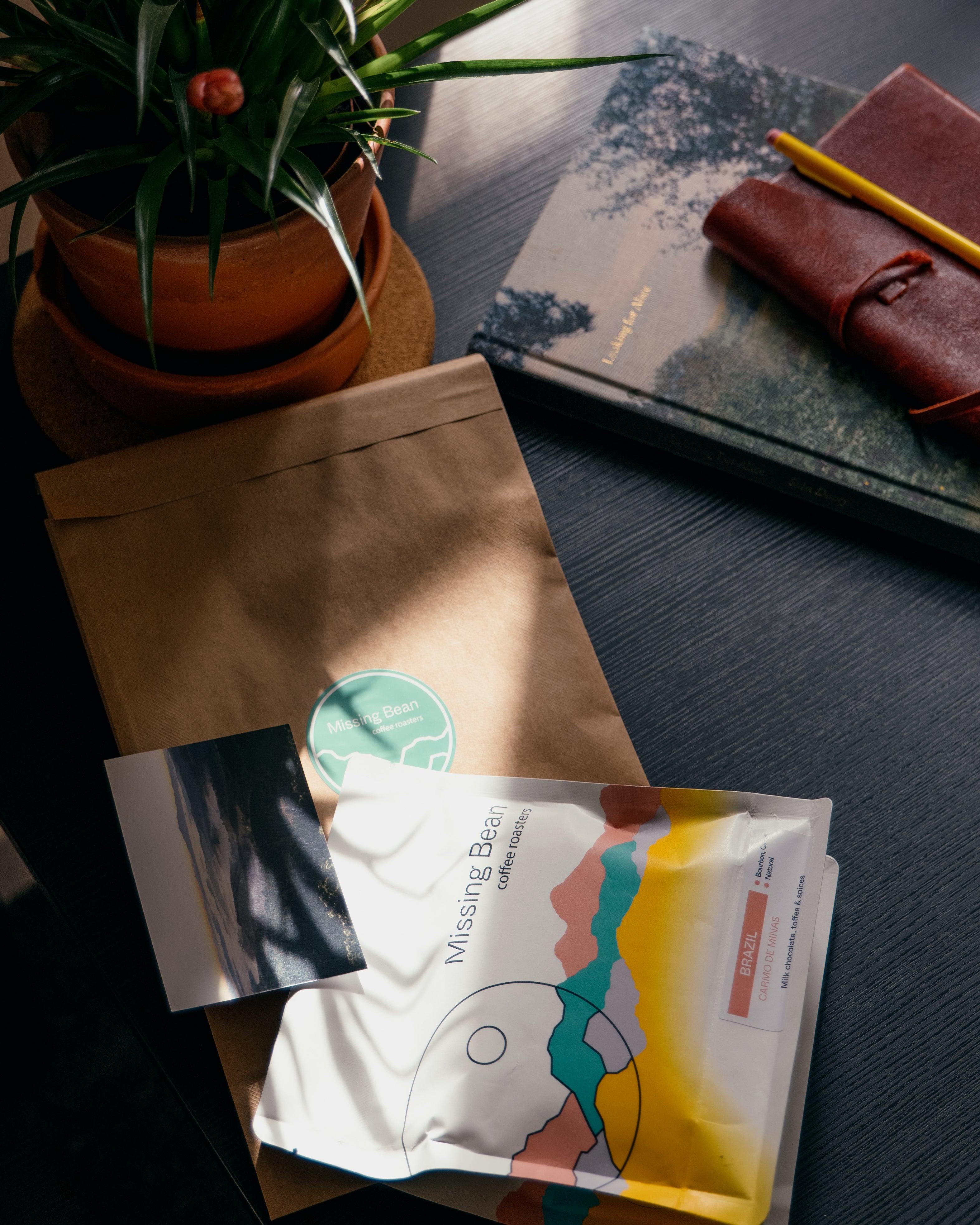 A colourful bag of Missing Bean's coffee beans is on a table along with information card, and brown recyclable envelope. There is a plant on the table, along with a book, notebook and pencil.