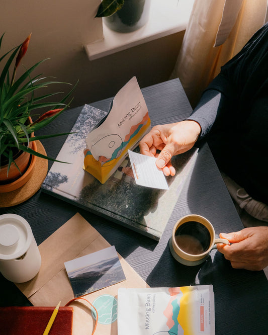 Person reading a coffee information card while holding a mug of black coffee on a table with a "Missing Bean Coffee Roasters" bag, a notebook, and a plant.