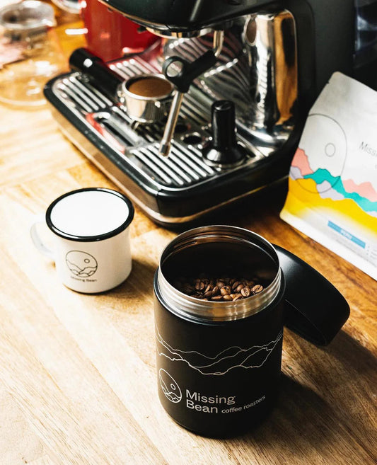 Open canister filled with coffee beans, a branded "Missing Bean Coffee Roasters" mug, and an espresso machine on a wooden countertop.