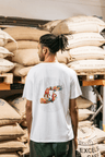A person in front of stacked burlap sacks of coffee beans. They are wearing a white Missing bean X Brian Gray T-Shirt featuring a colorful graphic design on the back, depicting two hands spelling out MB which are detailed with a range of clever, original, sketches to signify the journey of a coffee bean.