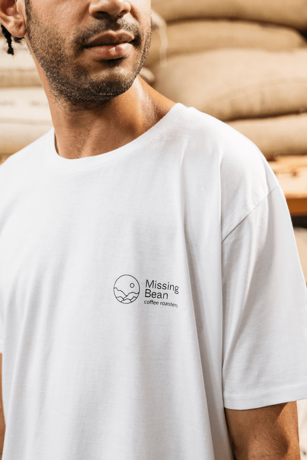 A man models a white Missing bean X Brian Gray T-Shirt. The front of the T-shirt has a black Missing Bean coffee roasters logo.